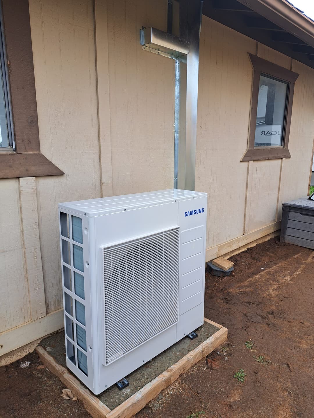 Samsung outdoor HVAC unit installed beside a wooden building with clear sky above.