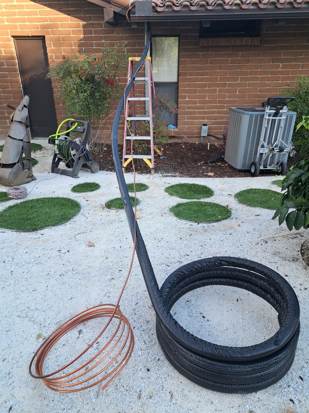 Coiled black wire and copper tubing in a garden setting with lawn, ladder, and tools.