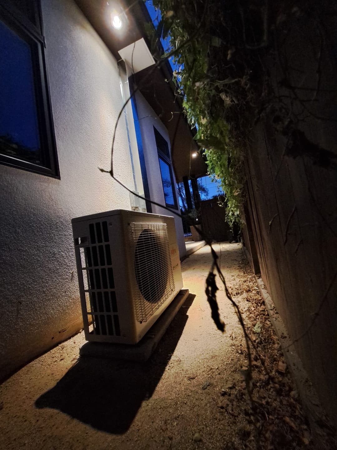 Exterior view of an air conditioning unit beside a house at dusk, surrounded by foliage.
