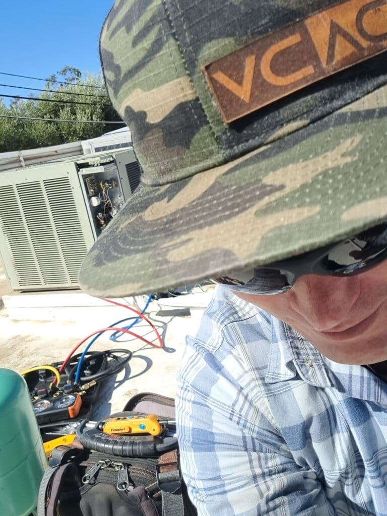 Technician in a camouflage cap working on HVAC equipment with tools and equipment nearby.